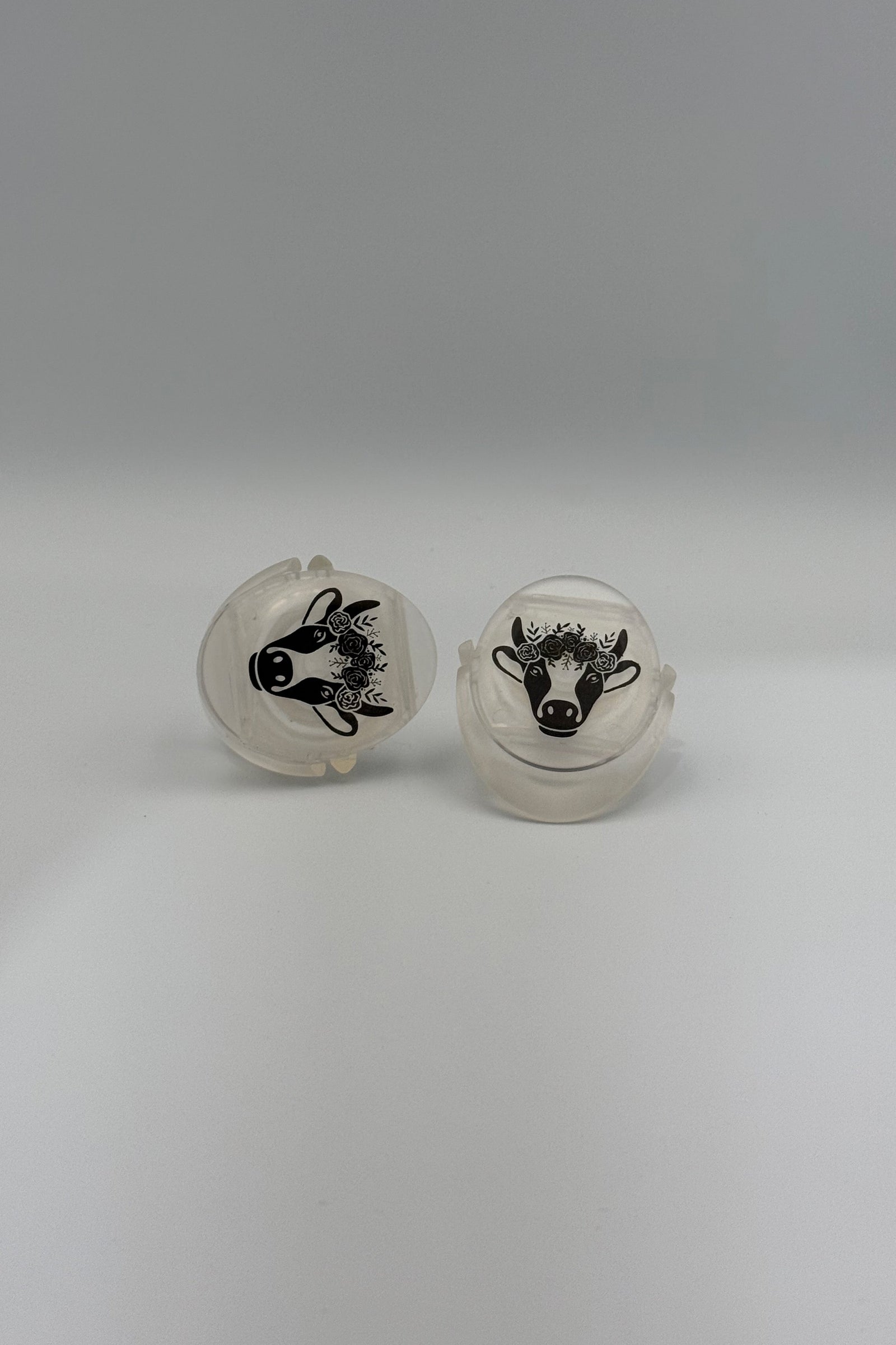 A pair of round, off-white plastic or acrylic novelty plugs with black animal illustrations, one depicting a cow and the other a rabbit, set against a plain gray background.